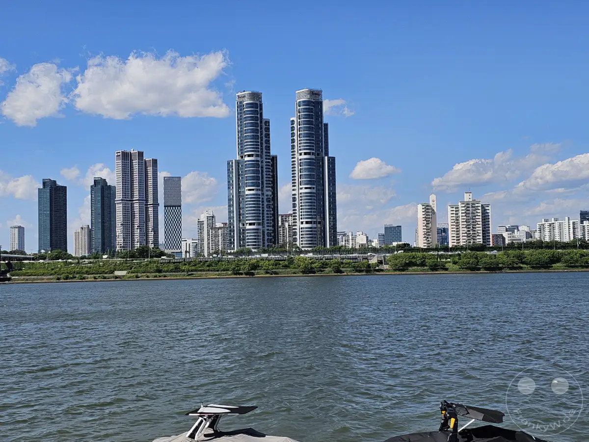 Die beeindruckende Skyline von Seoul spiegelt sich im ruhigen Wasser des Hangang-Flusses unter einem strahlend blauen Himmel mit vereinzelten Wolken. Moderne Wolkenkratzer und Hochhäuser säumen das Ufer und zeugen von der dynamischen Urbanität der südkoreanischen Hauptstadt. Die Szenerie vermittelt eine friedliche und klare Atmosphäre an einem sonnigen Tag.
