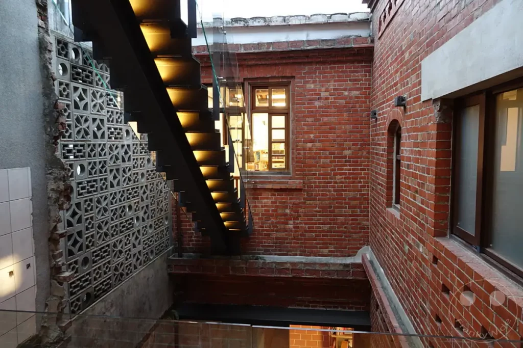 Eine Außentreppe aus schwarzem Metall mit integrierter Beleuchtung windet sich an der Backsteinwand eines historischen Gebäudes in Taipeh, Taiwan. Die interessante Musterung der Betonsteine und die roten Ziegelwände verleihen der Szene eine architektonische und nostalgische Atmosphäre. Durch ein Fenster im Obergeschoss dringt warmes Licht nach außen und deutet auf das Leben im Inneren hin.