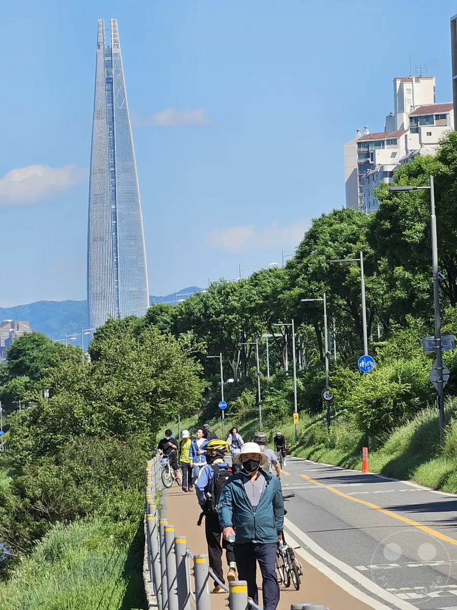 Menschen genießen einen sonnigen Tag auf einem Rad- und Fußweg entlang des Hangang-Flusses in Seoul, Südkorea. Im Hintergrund ragt die beeindruckende Lotte World Tower Skyline empor, umgeben von üppigem Grün. Die Szene strahlt eine entspannte und aktive Atmosphäre aus, während Radfahrer und Spaziergänger die urbane Natur genießen.