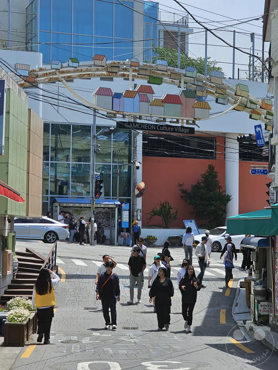 Touristen schlendern durch eine lebhafte Gasse im Culture Village Gamcheon in Busan, Südkorea. Die bunte Architektur und die kunstvollen Dekorationen über der Straße schaffen eine einzigartige Atmosphäre. Viele Menschen genießen den sonnigen Tag und erkunden die malerischen Straßen des Dorfes.