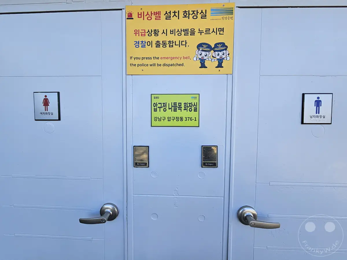 Diese Aufnahme zeigt die Eingänge von getrennten Toiletten am Hangang in Seoul, Südkorea, jeweils mit einem Schild für Damen und Herren. Eine über dem mittleren Eingang angebrachte Tafel informiert über einen Notrufknopf zur Verständigung der Polizei. Die Szene vermittelt einen informativen und gleichzeitig freundlichen Eindruck, verstärkt durch die "Be Happy"-Aufkleber.