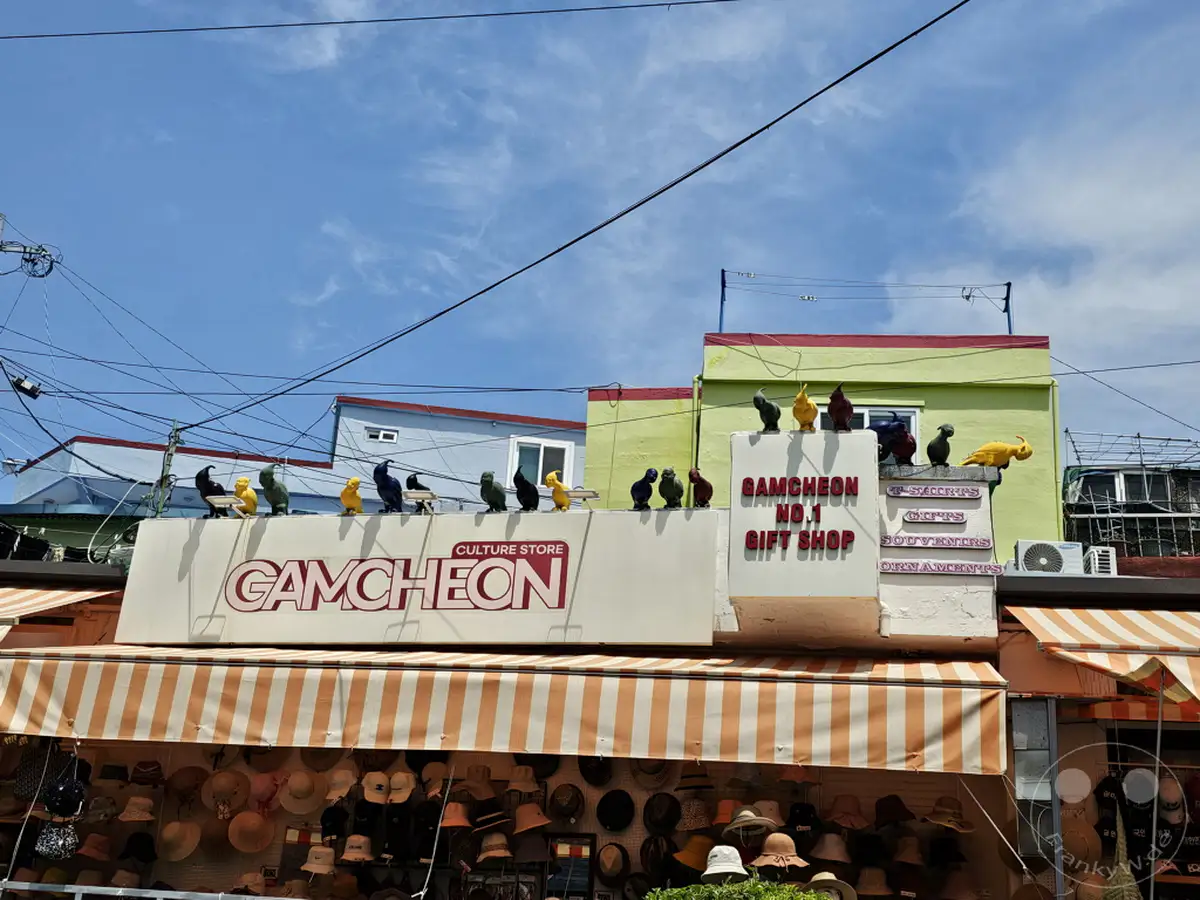 Ein bunter Souvenirshop im Culture Village Gamcheon in Busan, Südkorea, präsentiert sich unter einer gestreiften Markise. An der Fassade stehen dekorative Taubenfiguren, die auf einem Schild mit dem Aufdruck "GAMCHEON NO.1 GIFT SHOP" thronen. Die Szene versprüht eine lebhafte und touristische Atmosphäre unter einem blauen Himmel.