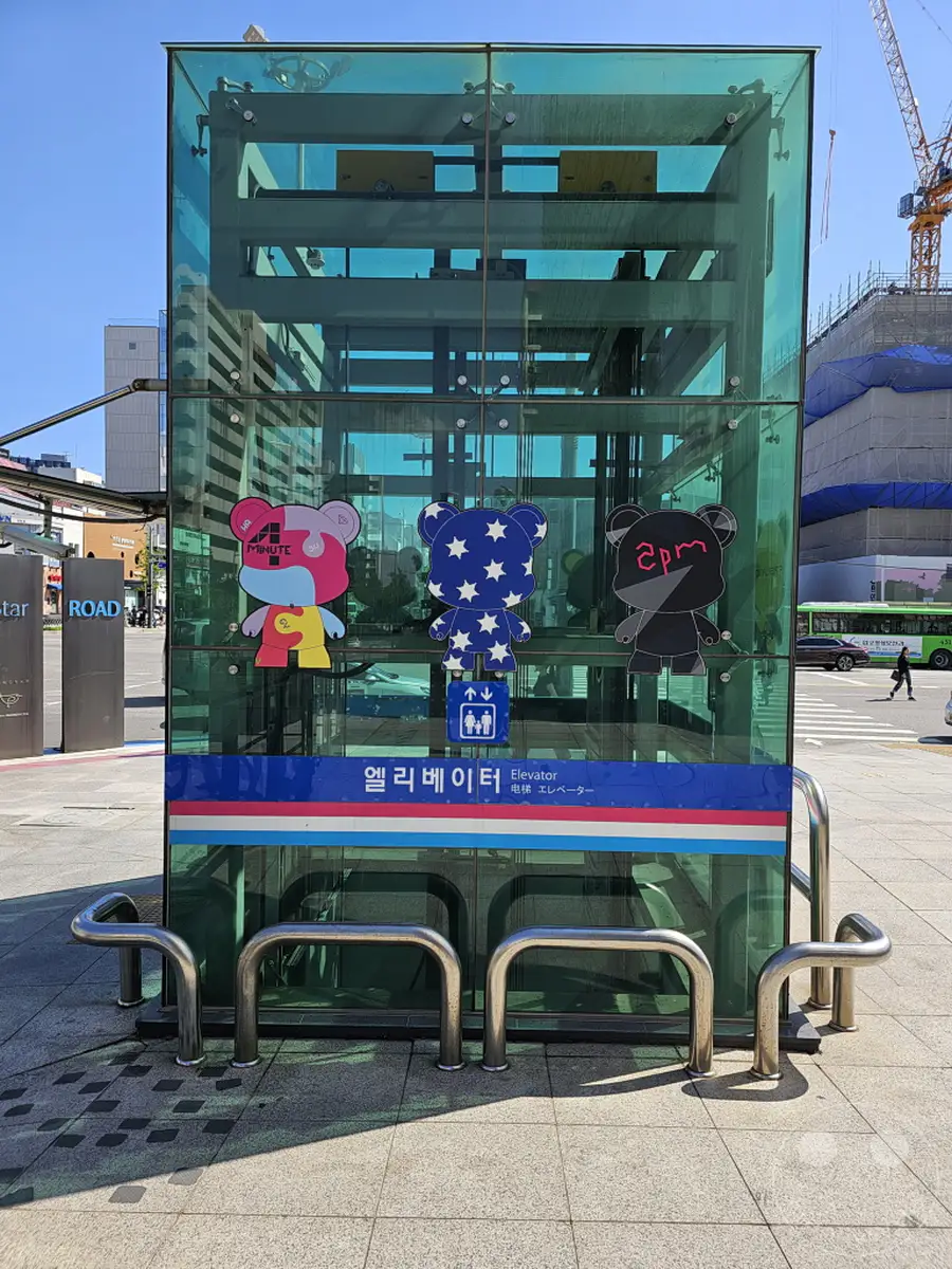 Ein moderner Glasaufzug steht auf einem gepflasterten Platz in Seoul, Südkorea, vor dem Hintergrund einer Baustelle und städtischer Gebäude. An der Glasfront sind bunte, sternenverzierte Bärenfiguren angebracht, die dem Aufzug eine verspielte und künstlerische Note verleihen. Die Szenerie deutet auf eine lebhafte und kreative urbane Umgebung hin, beleuchtet von hellem Sonnenschein.