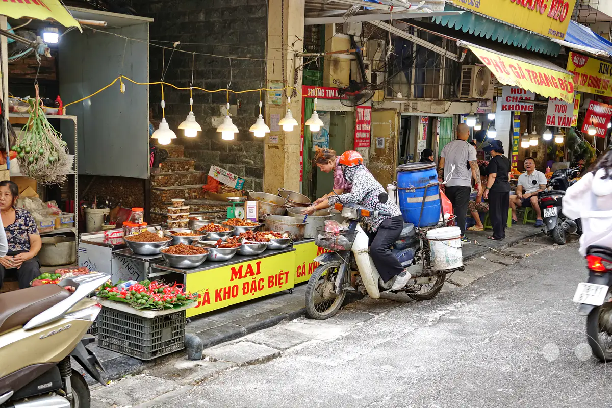 In Hanois lebendiger Altstadt, dem Old Quarter, bieten Händler auf einem geschäftigen Markt an einer Straße ihre Waren an. Dampfende Töpfe mit Speisen und frisches Gemüse sind auf den Tischen und an den Ständen ausgebreitet, während Menschen auf Motorrollern und zu Fuß den Alltag bewältigen. Die Szene fängt die geschäftige Atmosphäre und das kulinarische Treiben des vietnamesischen Alltags ein.