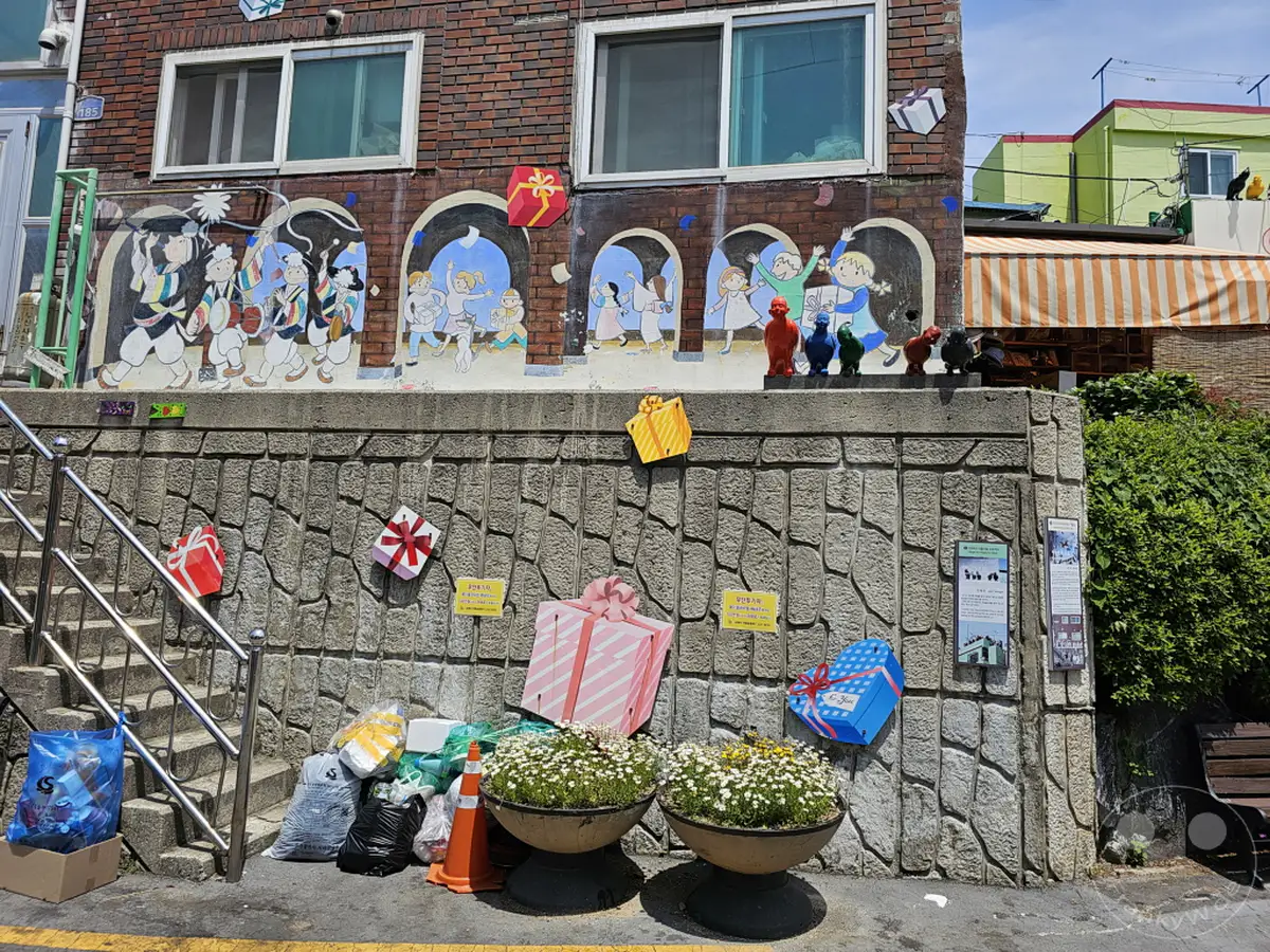 Die farbenfrohe Wandmalerei im Culture Village Gamcheon in Südkorea stellt fröhliche Szenen dar, die mit bunten Geschenken dekoriert sind. Ein steiler Treppenaufgang führt zu dem bemalten Gebäude, während zu beiden Seiten Dekorationen und Pflanzenarrangements zu sehen sind. Die Szenerie vermittelt eine festliche und künstlerische Atmosphäre in Busan.