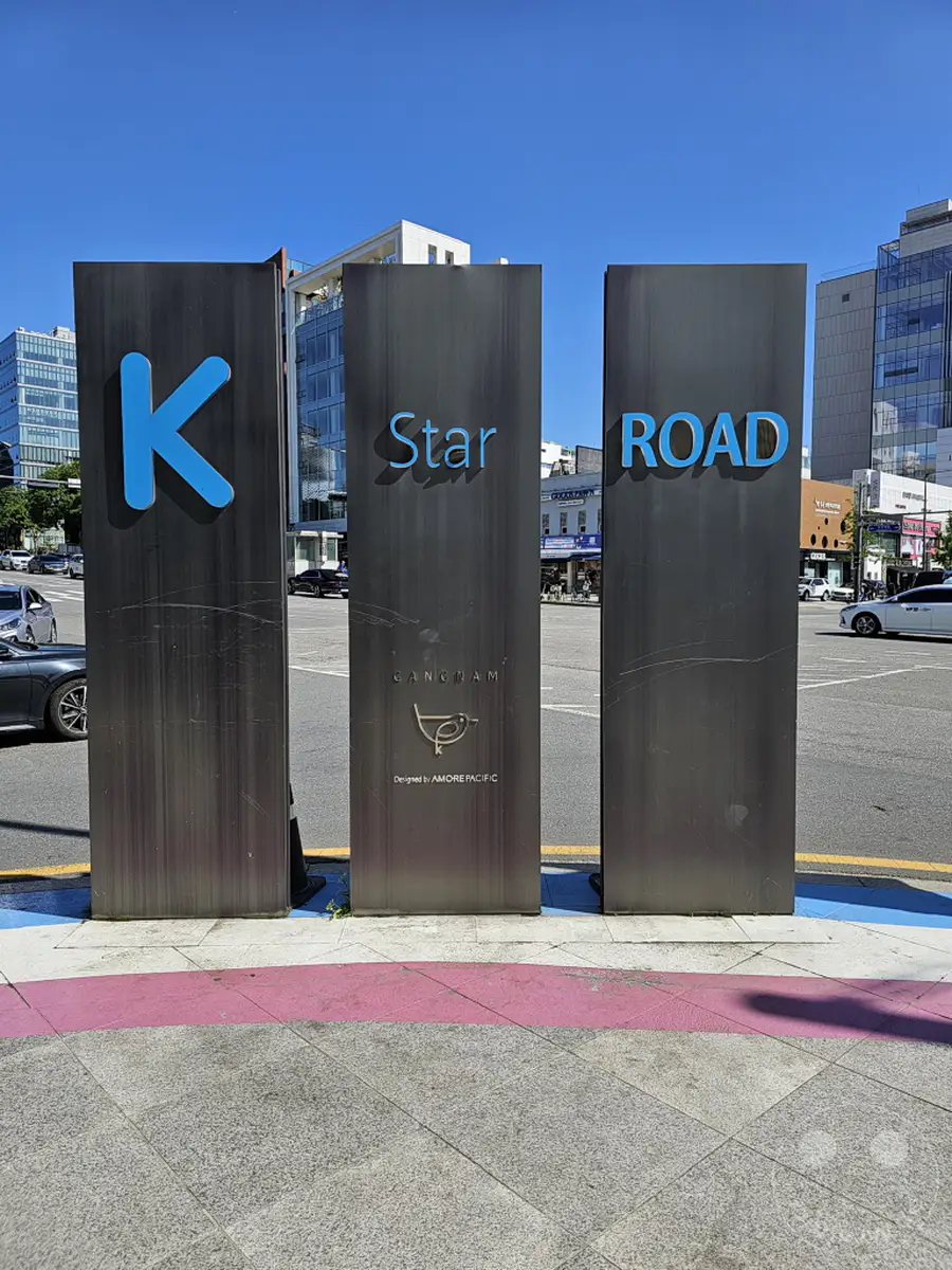 Drei schlanke, dunkle Metallstelen stehen nebeneinander auf einem Gehweg in Seoul. Die Stelen tragen die Aufschrift „K Star Road“, wobei der Buchstabe „K“ auf der ersten Stele in leuchtendem Blau hervorgehoben ist. Im Hintergrund sind moderne Gebäude und eine belebte Straße zu sehen, was auf die urbane Atmosphäre der Stadt hinweist.