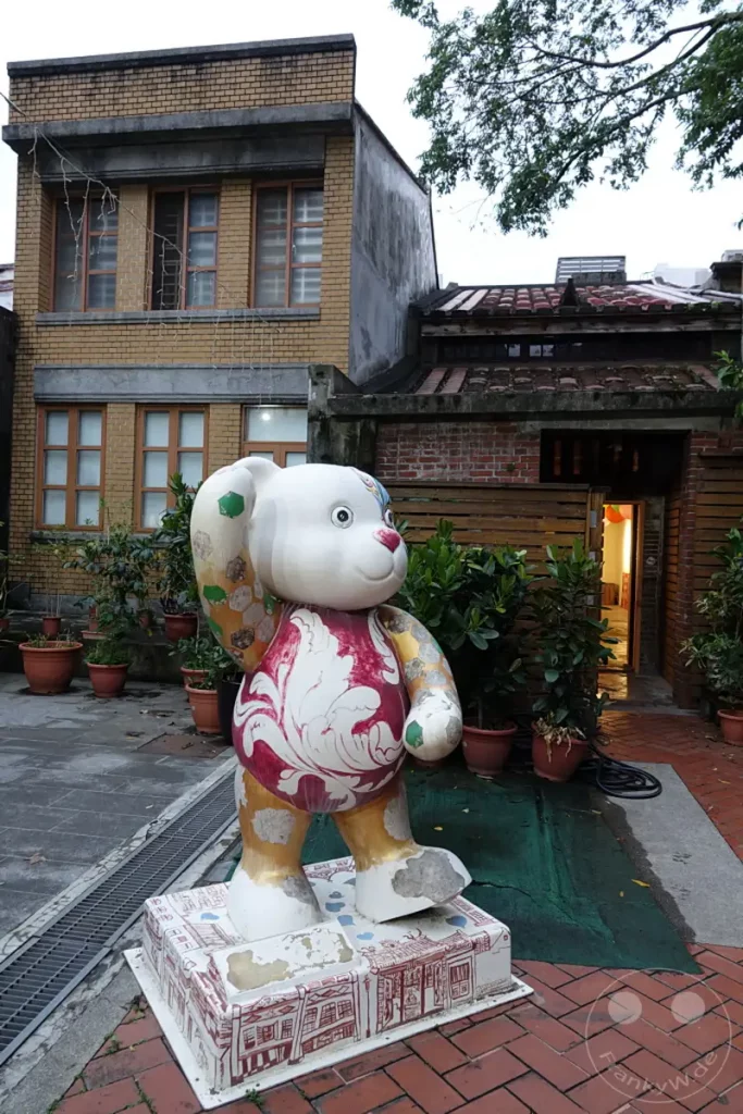 Eine große, bunte Bär-Statue steht auf einem Sockel vor einem Gebäude aus gelben Ziegeln in Taipeh, Taiwan. Im Hintergrund sind traditionelle taiwanische Dächer und eine hölzerne Fassade zu sehen, die eine nostalgische Atmosphäre schaffen. Die Szene vermittelt einen Eindruck von der Mischung aus moderner Kunst und historischem Erbe, die in der Dihua Street zu finden ist.