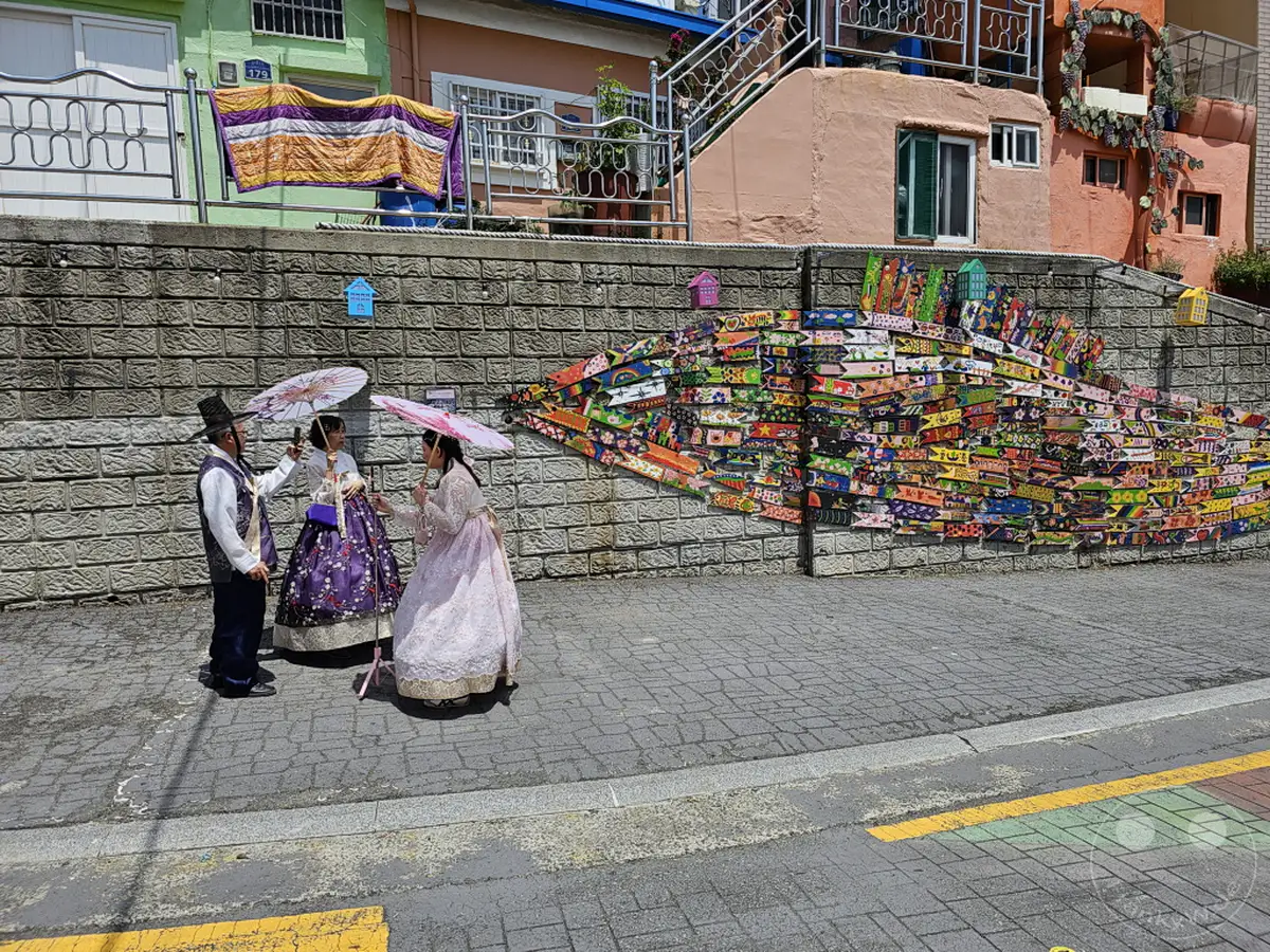 Drei Personen in traditionellen Hanbok-Kleidern posieren mit Schirmen vor einer kunstvollen Wandmalerei in Südkoreas Busan. Das farbenfrohe Wandbild, das eine Fischform nachbildet, schmückt eine Mauer im beliebten Culture Village Gamcheon. Die Szene strahlt eine Atmosphäre kultureller Wertschätzung und künstlerischer Kreativität aus.