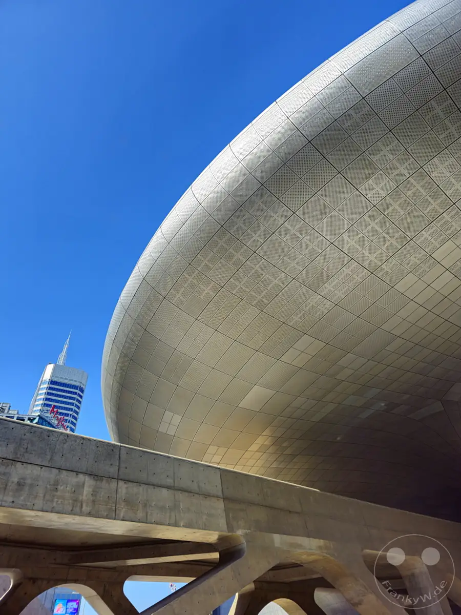 Eine futuristisch anmutende Fassade aus metallischen Paneelen erhebt sich unter einem strahlend blauen Himmel in Seoul, Südkorea. Im Vordergrund unterstreicht eine Betonbrücke mit markanten Bögen die avantgardistische Ästhetik des Dongdaemun Design Plaza (DDP). Das Bild fängt die architektonische Kühnheit und die klare Linienführung moderner Baukunst ein.