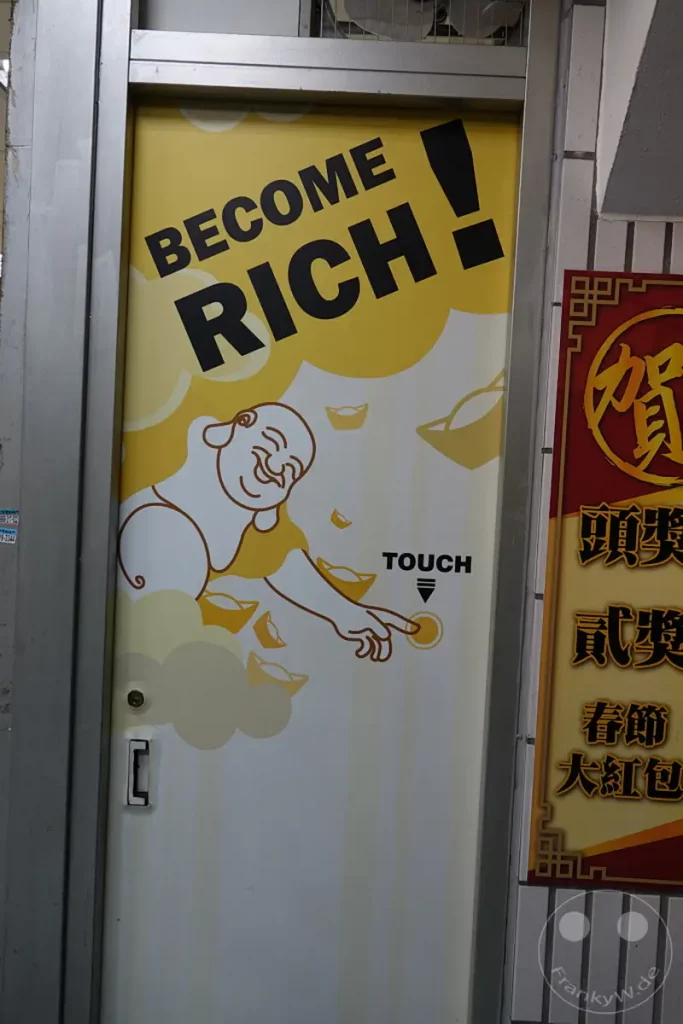 In Taipeh, Taiwan, auf der Dihua Street, ziert ein bunter Aufkleber eine Tür mit der Aufforderung "BECOME RICH!". Ein lächelnder Glücksbuddha zeigt auf einen Touch-Button, umgeben von symbolischen Goldbarren, was eine fröhliche und verheißungsvolle Atmosphäre schafft.