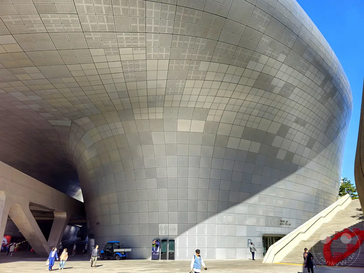 Das Dongdaemun Design Plaza (DDP) in Seoul, Südkorea, präsentiert sich in seiner vollen futuristischen Pracht unter strahlend blauem Himmel. Die gewölbte, metallische Fassade des Gebäudes, charakterisiert durch seine einzigartige Form und die perforierte Oberfläche, wirft markante Schatten auf den Platz. Menschen bewegen sich entspannt in der offenen und modernen Umgebung, während eine Treppe zu einem höheren Zugangspunkt führt und die geschäftige Atmosphäre einer lebendigen Metropole unterstreicht.