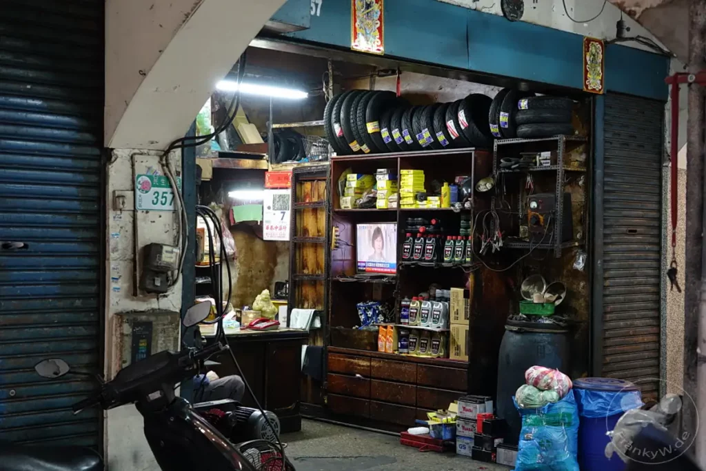 Eine kleine Motorradwerkstatt in Taipeh, Taiwan, in der Dihua Street, ist bei Nacht beleuchtet. Reifen und Ersatzteile stapeln sich in einem Regal, während eine flackernde TV-Anzeige im Laden läuft. Die Szene vermittelt eine geschäftige und authentische Atmosphäre eines lokalen Geschäfts im städtischen Umfeld.