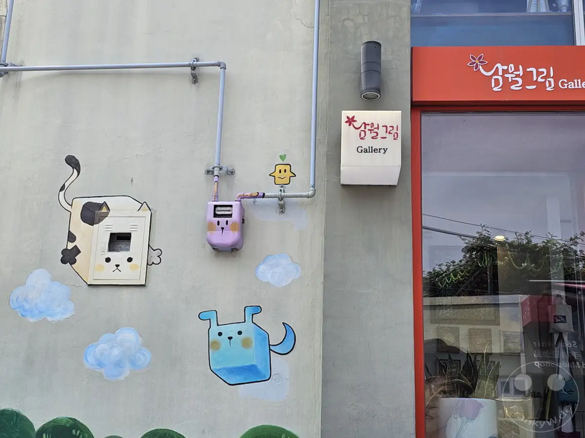 In Busan, Südkorea, ziert eine bunte Wandmalerei die Fassade eines Gebäudes, das Teil des berühmten Kulturdorfes Gamcheon ist. Die verspielten Charaktere und Wolkenbilder schaffen eine fröhliche und künstlerische Atmosphäre. Ein Schild mit der Aufschrift "Gallery" und koreanischer Schrift deutet auf eine Galerie hin, die diese kreative Umgebung ergänzt.