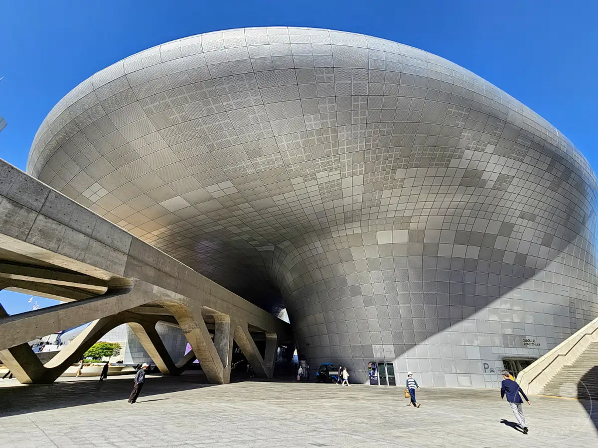 Der Dongdaemun Design Plaza (DDP) in Seoul, Südkorea, präsentiert sich unter strahlend blauem Himmel in seiner futuristischen Form. Die geschwungene Metallfassade wirft scharfe Schatten auf den gepflasterten Platz, auf dem einige Personen unterwegs sind. Ein stählerner Fußgängerweg überspannt den Außenbereich und unterstreicht die avantgardistische Ästhetik des Gebäudes.