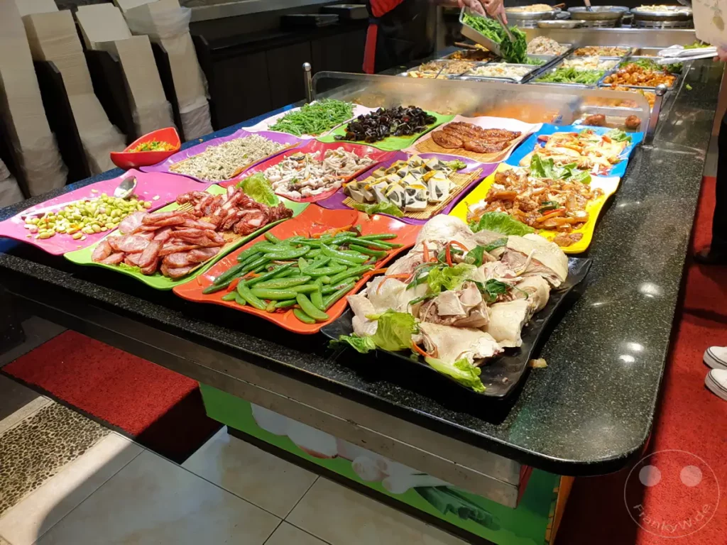 Ein vielfältiges Buffet mit zahlreichen asiatischen Gerichten wird in Taipeh, Taiwan, präsentiert. Die Auswahl reicht von deftigen Fleischgerichten über frisches Gemüse bis hin zu dampfgegarten Speisen, die zum Probieren einladen. Die Szene strahlt eine geschäftige und kulinarische Atmosphäre aus, typisch für die Märkte und Essensstände der Dihua Street.