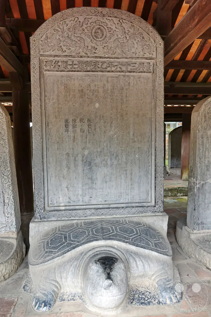 Eine steinerne Stele mit Schriftzeichen, getragen von einer Schildkröte, steht im Tempel der Literatur in Hanoi, Vietnam. Die stoische Haltung der Schildkröte und die kunstvollen Gravuren der Stele strahlen eine Aura von Geschichte und Weisheit aus. Diese Skulptur ist ein zentrales Element der Anlage und verkörpert die tiefe kulturelle Bedeutung des Ortes.