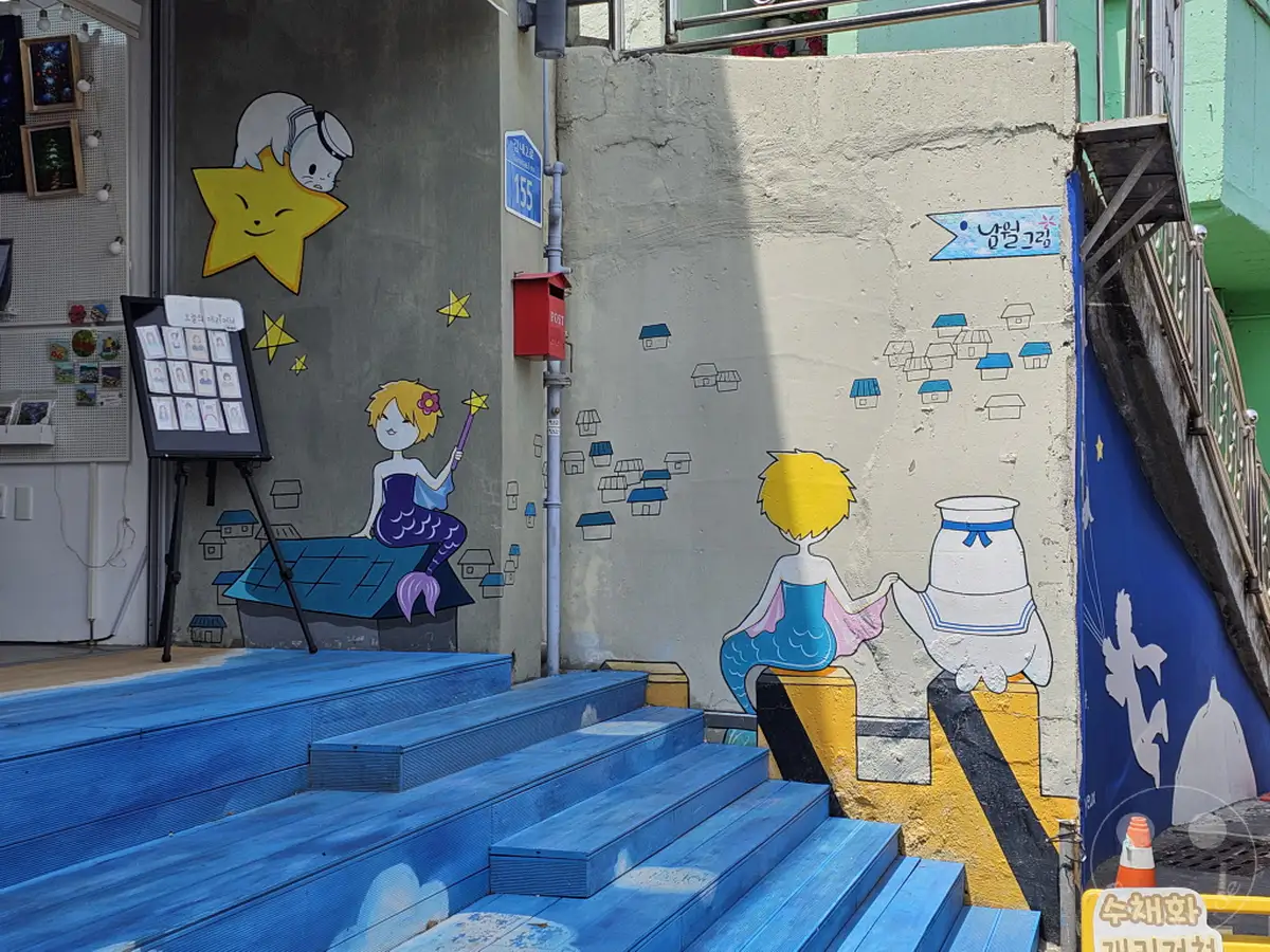 In Südkorea, genauer gesagt in Busan im Culture Village Gamcheon, zieren bunte Wandmalereien die Hausfassaden. Eine Meerjungfrau sitzt auf einem Dach neben einer kleinen Siedlung aus Häusern, während Sterne und eine Katze auf einem Stern auf dem grauen Putz abgebildet sind. Die leuchtend blauen Holzstufen führen zu diesem farbenfrohen Ort, der eine verspielte und fantasievolle Atmosphäre ausstrahlt.