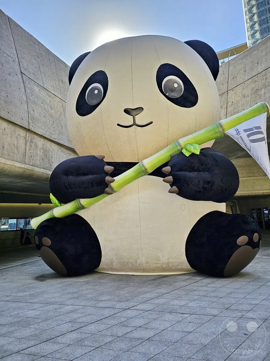 Ein riesiger aufblasbarer Panda hält einen Bambusstock mit einer Fahne des 'DDP 10th ANNIVERSARY' in Seoul, Südkorea. Die aufblasbare Skulptur ist vor dem Dongdaemun Design Plaza (DDP) aufgestellt, eine beeindruckende Installation im Freien. Die Sonne scheint hinter dem Kopf des Pandas hervor und wirft einen hellen Schein auf die Szene.