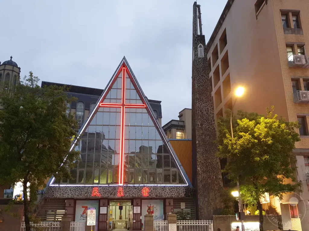 Eine moderne Kirche mit einem großen roten Kreuz aus Neonlicht dominiert die Fassade, während die umliegende Architektur und die Beleuchtung auf eine städtische Umgebung hinweisen. Die Szene vermittelt eine Mischung aus religiöser Symbolik und dem lebendigen Flair eines Stadtviertels wie dem Ningxia Nightmarket in Taipeh, Taiwan. Die Dämmerung oder Nacht deutet auf eine friedliche, aber auch geschäftige Atmosphäre hin.