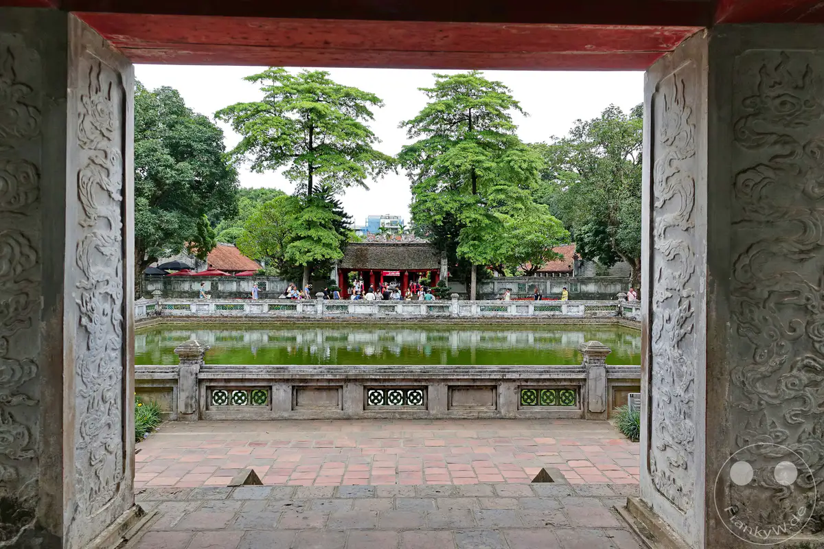 Der Blick durch einen verzierten Torbogen enthüllt den Literaturtempel in Hanoi, Vietnam, mit seinem ruhigen Teich und traditionellen Gebäuden. Mehrere Bäume mit üppig grünem Laub säumen den Teich, während Besucher entlang der Balustrade auf der gegenüberliegenden Seite flanieren. Die Szenerie strahlt eine friedliche Atmosphäre der Entdeckung und historischen Besinnung aus.