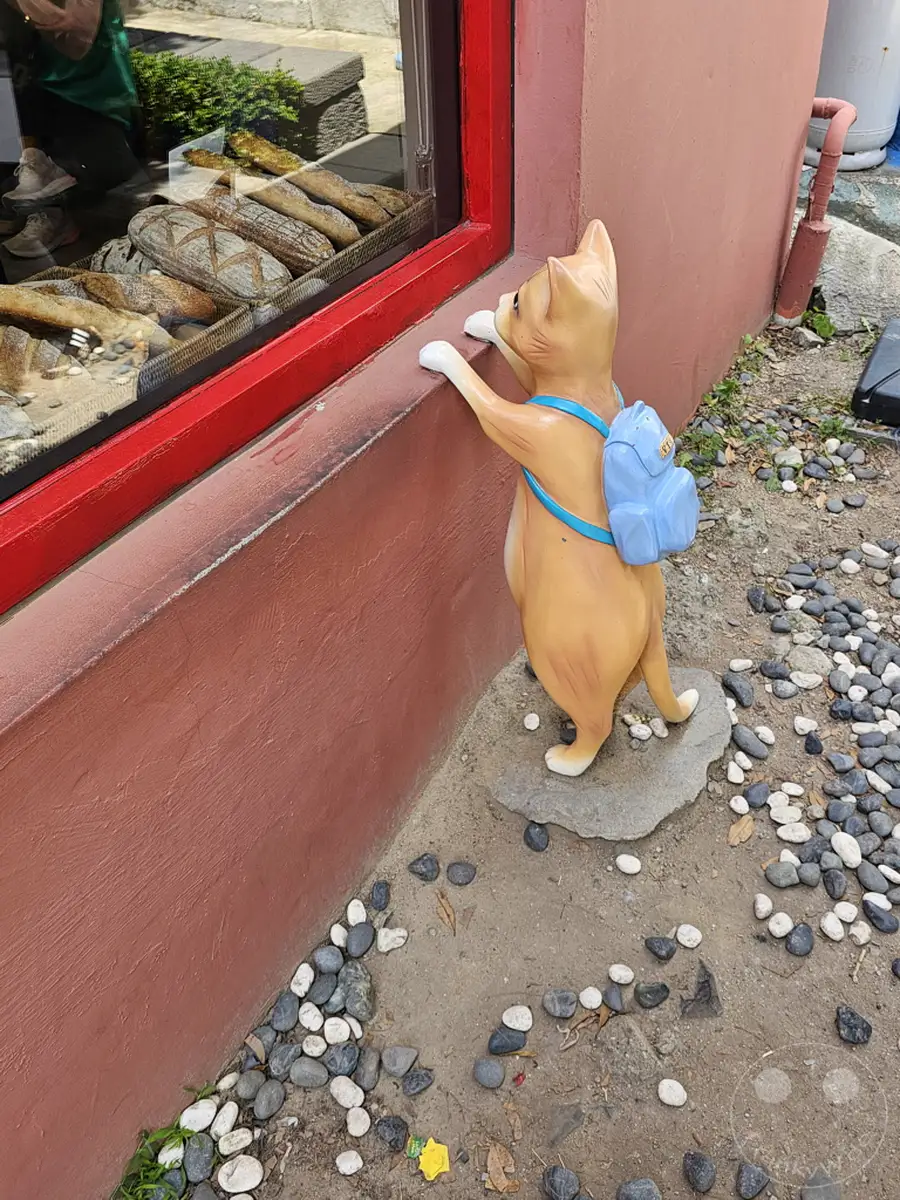 Eine skurrile Katzenskulptur mit Rucksack steht neugierig vor dem Fenster einer Bäckerei im Culture Village Gamcheon in Busan, Südkorea. Die Bäckerei zeigt eine verlockende Auswahl an frischen Broten, die durch das Fenster zu sehen sind. Die Szene vermittelt eine charmante und spielerische Atmosphäre, die typisch für das künstlerische Viertel ist.