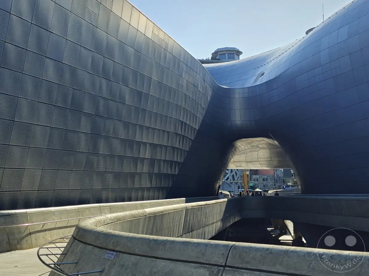 Das Dongdaemun Design Plaza (DDP) in Seoul, Südkorea, präsentiert sich mit seiner futuristischen, organischen Architektur unter einem klaren Himmel. Die geschwungenen Formen des Gebäudes und die umliegenden Betonstrukturen schaffen eine beeindruckende Kulisse für den öffentlichen Raum. Die Szene vermittelt ein Gefühl von Modernität und architektonischer Innovation.