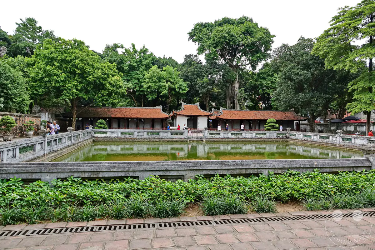 In Hanoi, Vietnam, steht das Literaturtempel-Komplex mit seiner historischen Architektur, umgeben von üppigen Grünpflanzen. Ein rechteckiges Wasserbecken spiegelt die roten Ziegeldächer und die Bäume wider und schafft eine ruhige Atmosphäre. Besucher schlendern entlang des Beckens und bewundern die traditionsreiche Anlage.