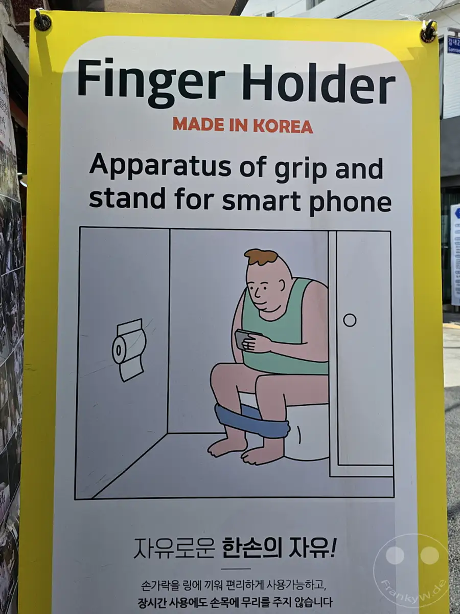 Eine gelbe Werbetafel in Südkorea, genauer gesagt im Busan Culture Village Gamcheon, wirbt für einen "Finger Holder" für Smartphones. Die Illustration zeigt humorvoll einen Mann auf der Toilette, der das Produkt benutzt. Der Slogan auf Koreanisch verspricht "freie Hände" und "komfortable Nutzung" auch bei langer Nutzung.