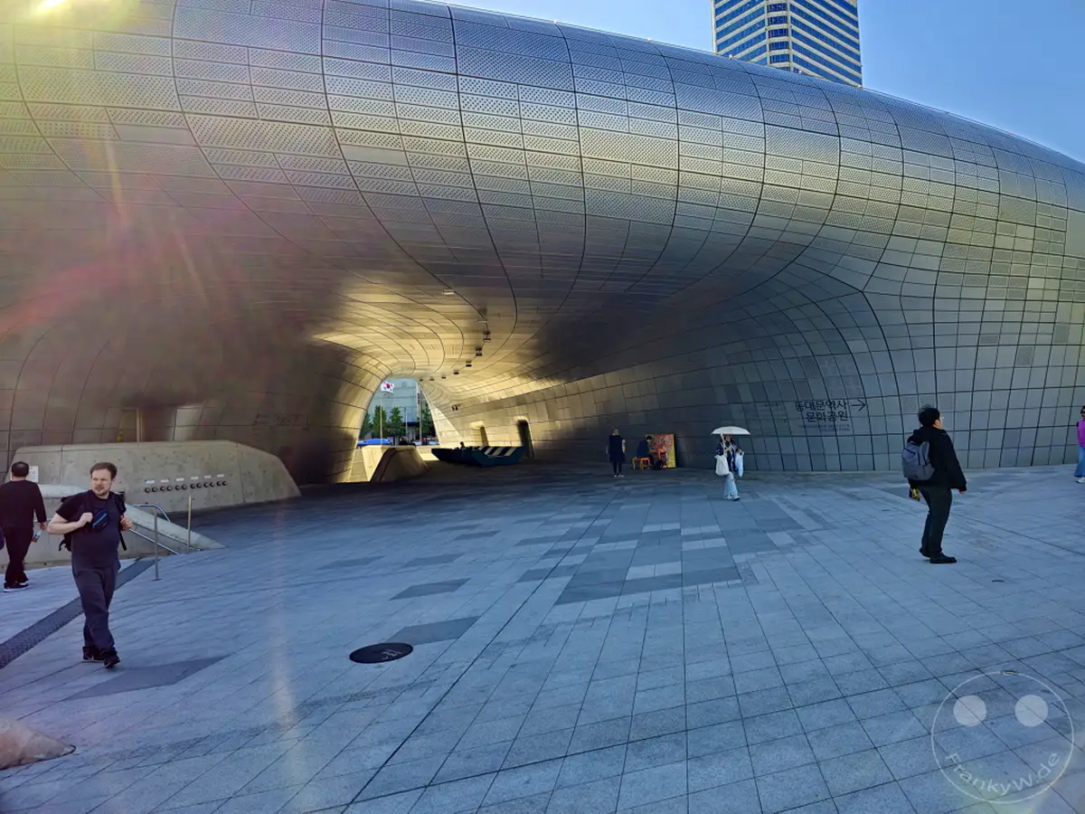 Die futuristische Architektur des Dongdaemun Design Plaza (DDP) in Seoul, Südkorea, erhebt sich vor einem klaren blauen Himmel. Menschen bewegen sich auf dem weitläufigen, gepflasterten Platz vor dem Gebäude, während die untergehende Sonne lange Schatten wirft und eine ruhige, aber belebte Atmosphäre schafft.