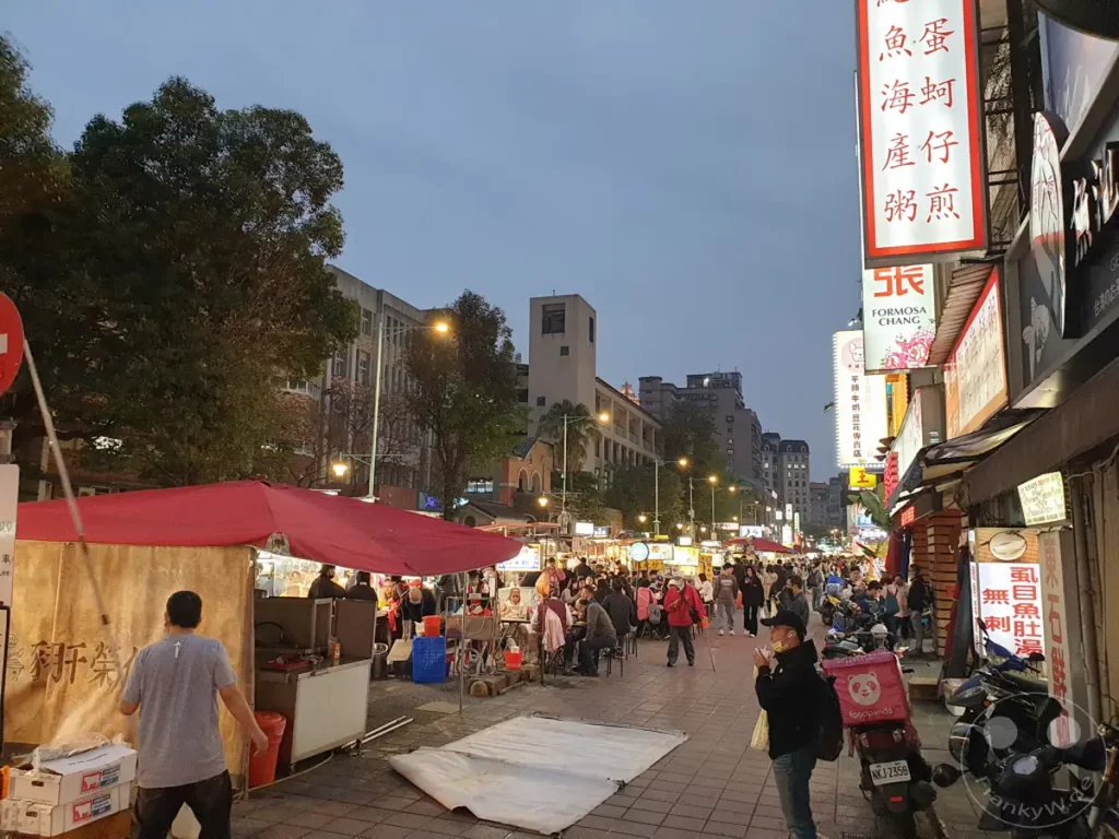 Die lebendige Atmosphäre des Ningxia Night Market in Taipeh, Taiwan, wird in dieser Aufnahme am Abend eingefangen. Garküchen säumen die Straße, während zahlreiche Besucher durch die Gassen schlendern und die kulinarischen Angebote genießen. Ein geschäftiges Treiben herrscht inmitten der beleuchteten Stände und der abendlichen Dämmerung.