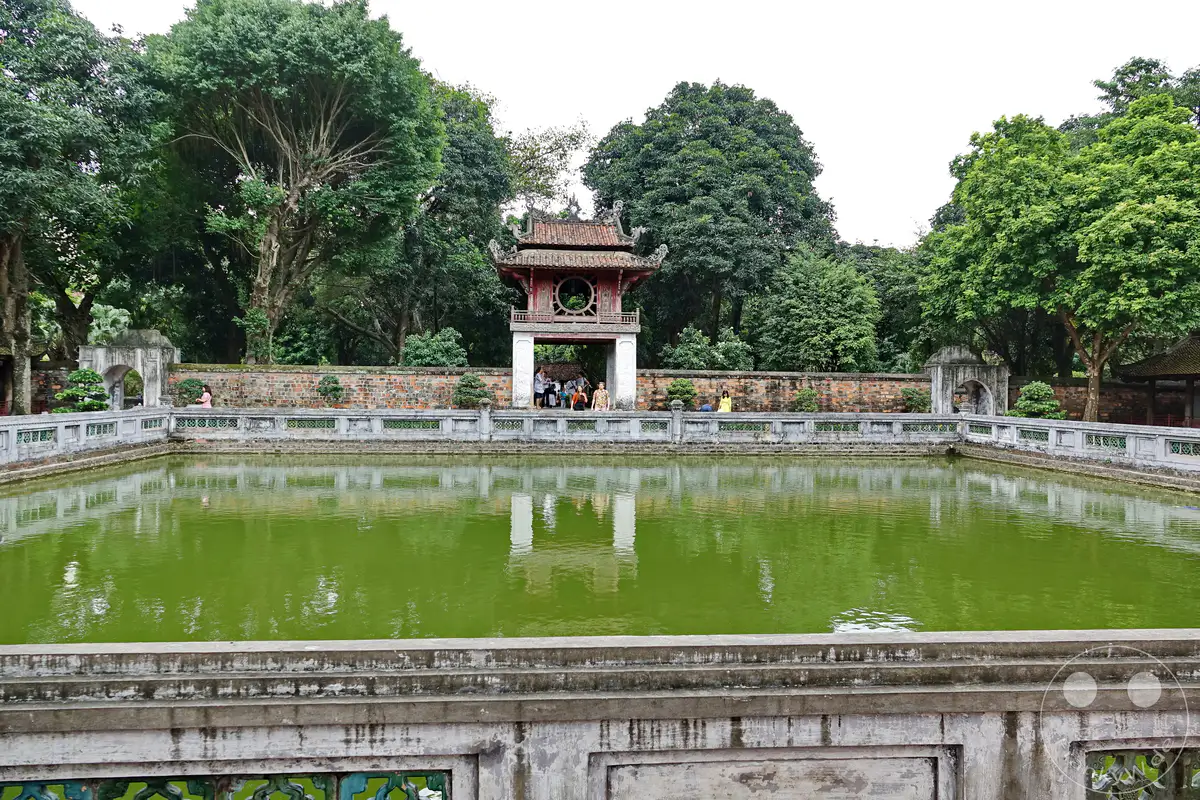 Der Tempel der Literatur in Hanoi, Vietnam, präsentiert eine historische Anlage mit einem ruhigen, smaragdgrünen Teich, der die Architektur und die üppige Vegetation widerspiegelt. Besucher spazieren entlang des Teichs und bewundern die traditionelle vietnamesische Architektur und die gepflegten Gärten. Die Szene strahlt eine friedliche und nachdenkliche Atmosphäre aus, die zum Entdecken einlädt.