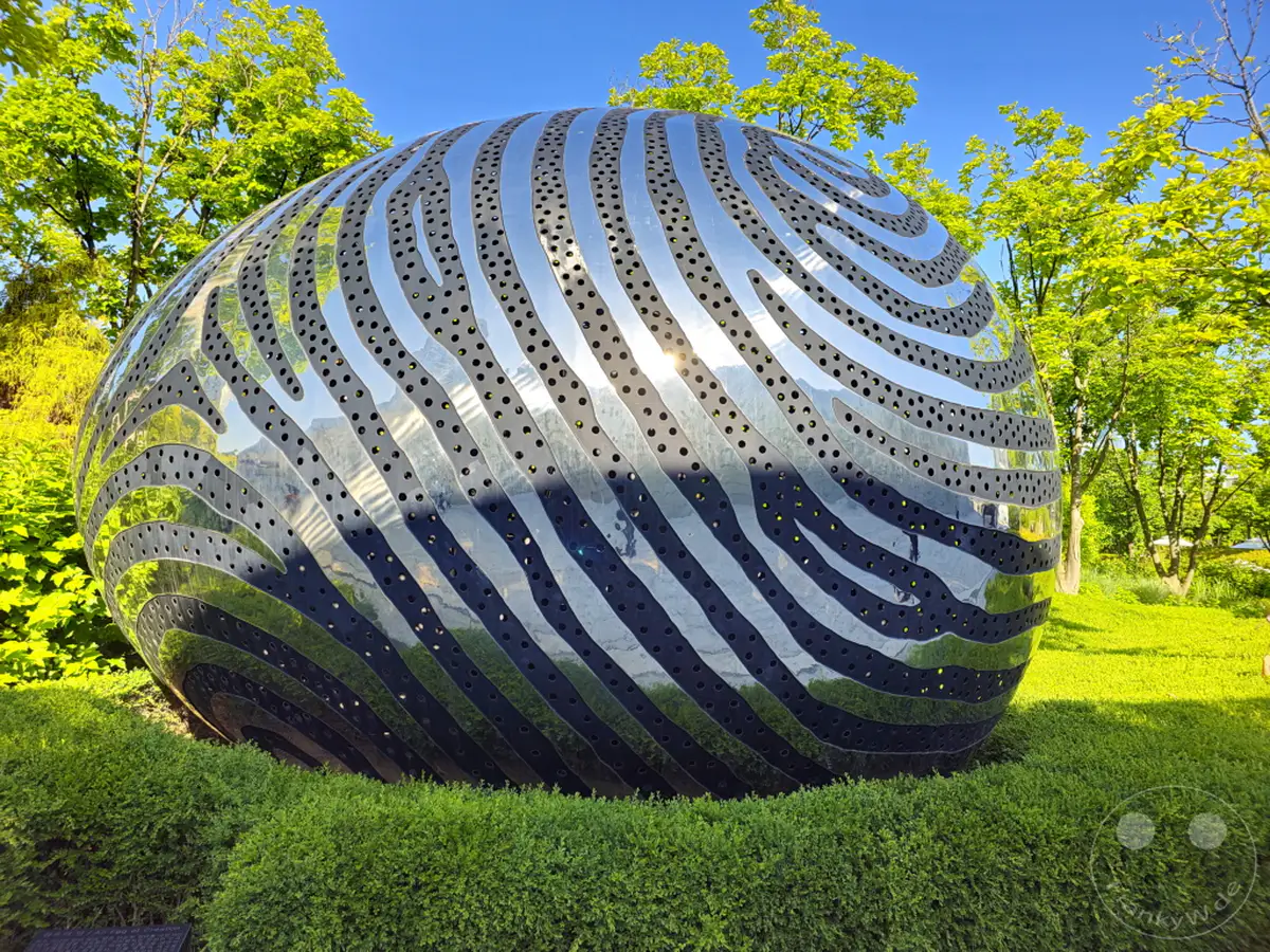 Eine futuristisch anmutende, kugelförmige Metallskulptur mit perforiertem Muster steht inmitten einer gepflegten Grünanlage im Dongdaemun Design Plaza (DDP) in Seoul, Südkorea. Die glänzende Oberfläche reflektiert die üppige grüne Umgebung und den strahlend blauen Himmel, was eine harmonische Verbindung von Kunst und Natur erzeugt. An einem sonnigen Tag entfaltet die Installation ihre beeindruckende Präsenz vor dem Hintergrund der modernen Architektur und der umliegenden Bäume.