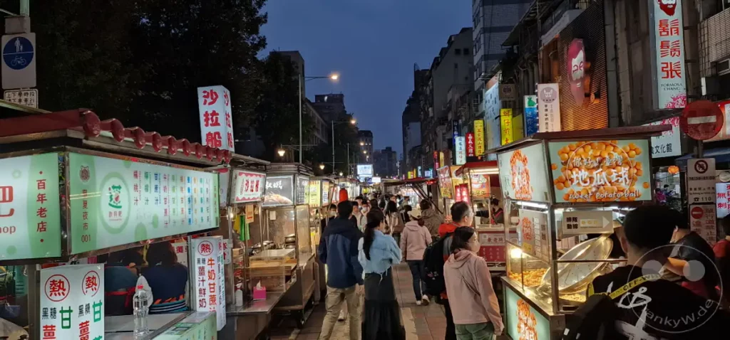 Eine belebte Nachtszene auf dem Ningxia Nightmarket in Taipeh, Taiwan, wo Essensstände mit bunten Lichtern und dampfenden Köstlichkeiten locken. Menschen schlendern durch die Gassen und genießen die lebendige Atmosphäre des nächtlichen Treibens. Viele Stände bieten traditionelles taiwanesisches Streetfood an, von süßen Kartoffelbällchen bis zu herzhaften Gerichten.