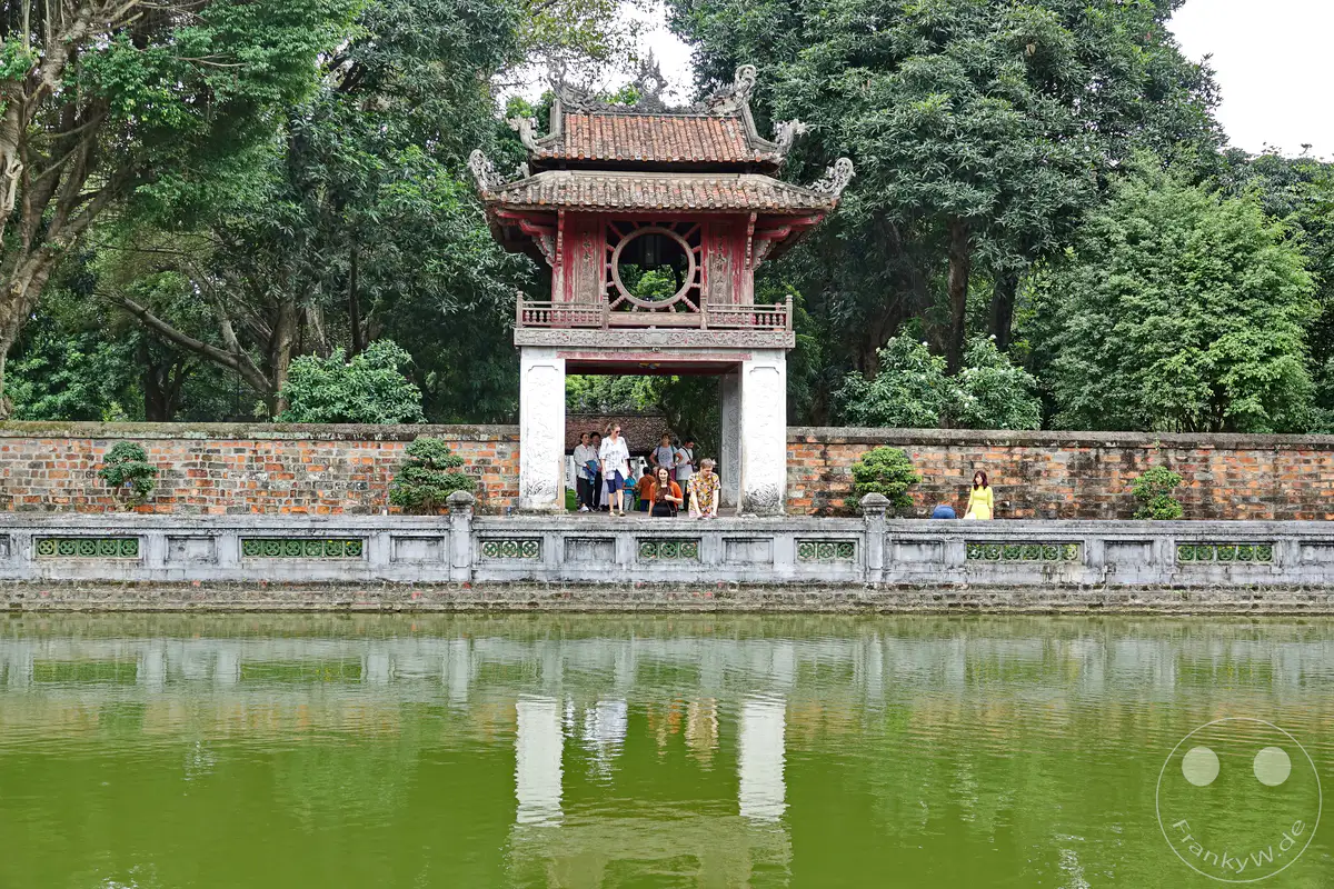 Der Literaturtempel in Hanoi, Vietnam, ist ein historisches Wahrzeichen mit einer traditionellen Pagode und einem ruhigen Teich. Besucher spazieren entlang der Mauer, während üppige Bäume die Szenerie umgeben und eine friedliche Atmosphäre schaffen.