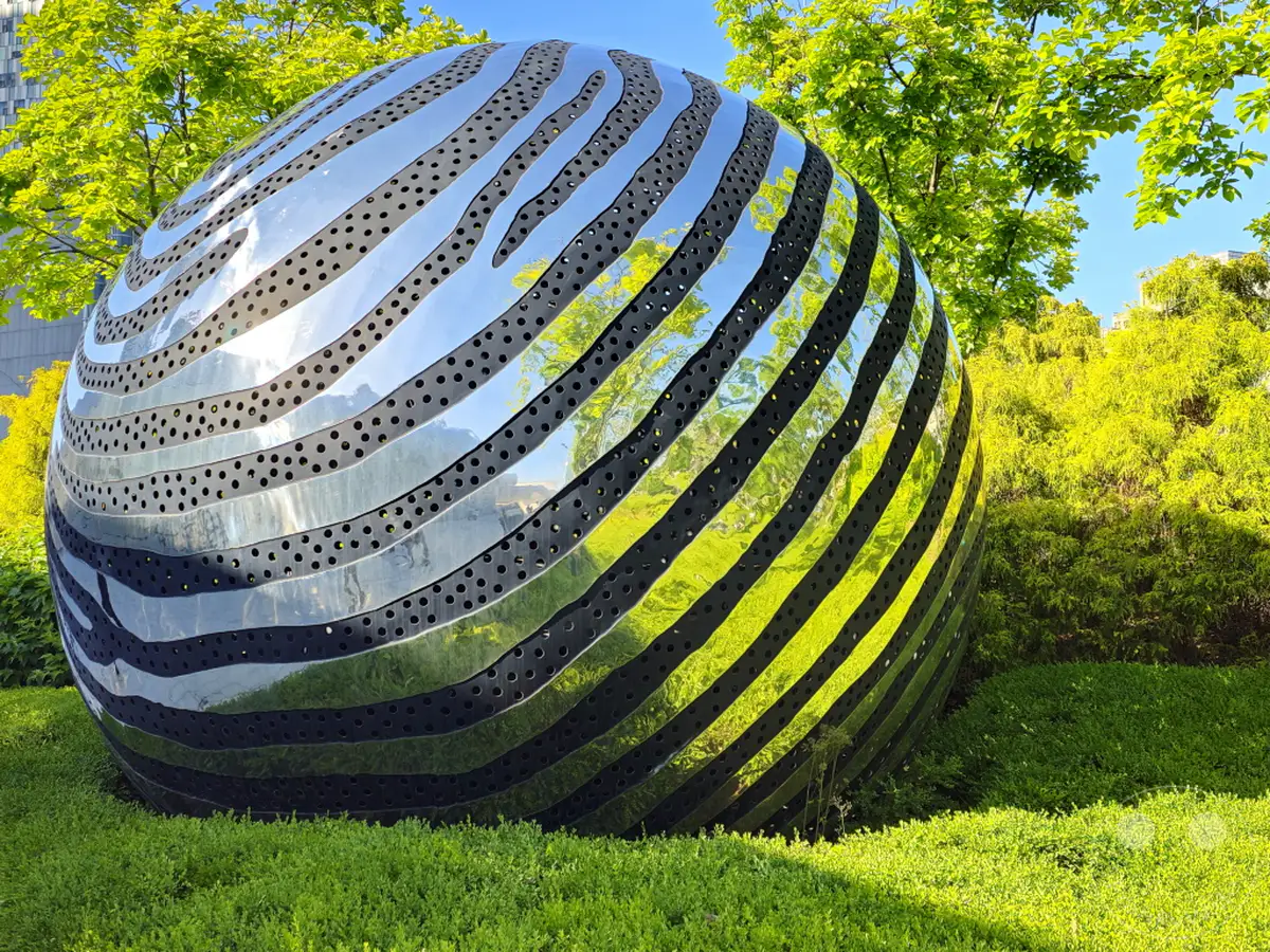 Eine beeindruckende, kugelförmige Metallskulptur mit einem Muster aus sich schlängelnden Streifen und zahlreichen kleinen Löchern steht in einer gepflegten Parkanlage im Dongdaemun Design Plaza (DDP) in Seoul, Südkorea. Das hochglanzpolierte Äußere der Skulptur reflektiert die üppig grüne Umgebung und den strahlend blauen Himmel, was eine moderne und zugleich naturverbundene Atmosphäre schafft. Sonneneinstrahlung wirft Licht und Schatten auf die kunstvolle Installation und die umliegenden Bäume und Büsche.