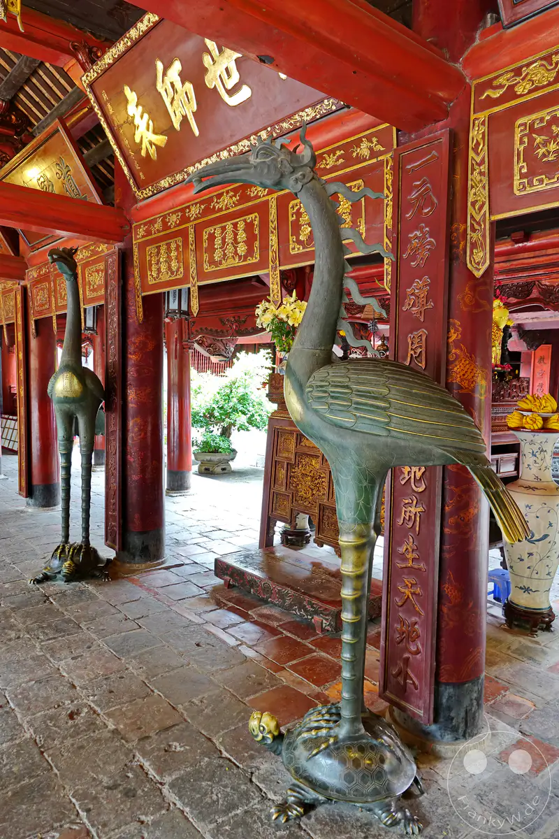 Zwei bronzene Kranich-Statuen, jede auf einer Schildkröte stehend, sind im Tempel der Literatur in Hanoi, Vietnam, aufgestellt. Die Säulen und Wände sind mit traditionellen roten und goldenen Verzierungen geschmückt, was eine Atmosphäre von Ehrfurcht und historischer Bedeutung schafft. Das historische Gebäude mit seinen filigranen Details lädt zum Entdecken der vietnamesischen Kultur ein.