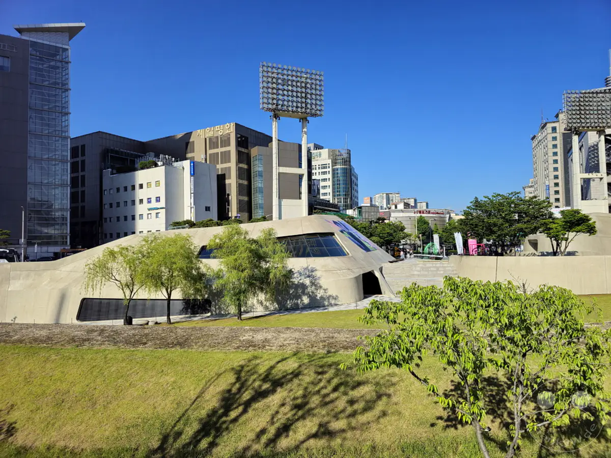 Diese Aufnahme von Seoul, Südkorea, zeigt das beeindruckende Dongdaemun Design Plaza (DDP) an einem sonnigen Tag. Moderne Gebäude und grüne Parkflächen bilden einen spannenden Kontrast zur futuristischen Architektur des DDP. Die klare blaue Himmel und das satte Grün der Vegetation schaffen eine lebendige und erfrischende Atmosphäre in der städtischen Umgebung.