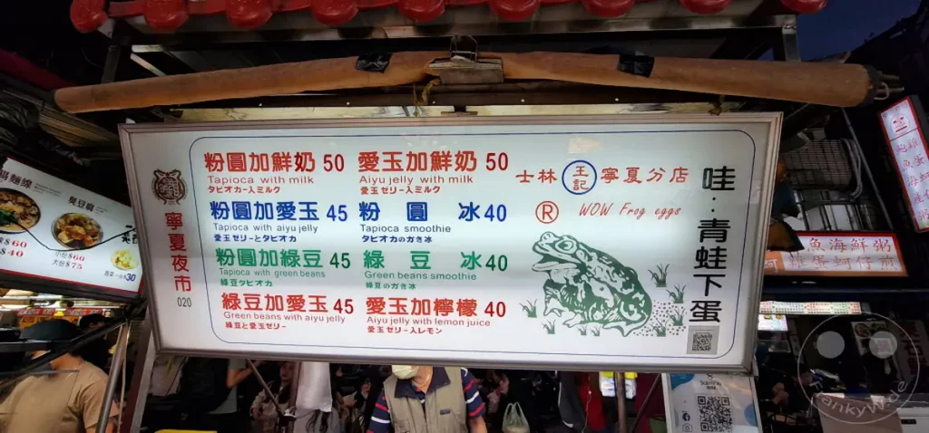 In Taipeh, Taiwan, präsentiert der Ningxia Nightmarket eine verlockende Auswahl an Streetfood-Angeboten. Leuchtende Schilder mit chinesischen und englischen Menüs deuten auf süße und erfrischende Getränke hin, während ein Cartoon-Frosch für die "Wow Frog Eggs" wirbt. Die lebhafte Atmosphäre eines nächtlichen Marktes ist deutlich spürbar.