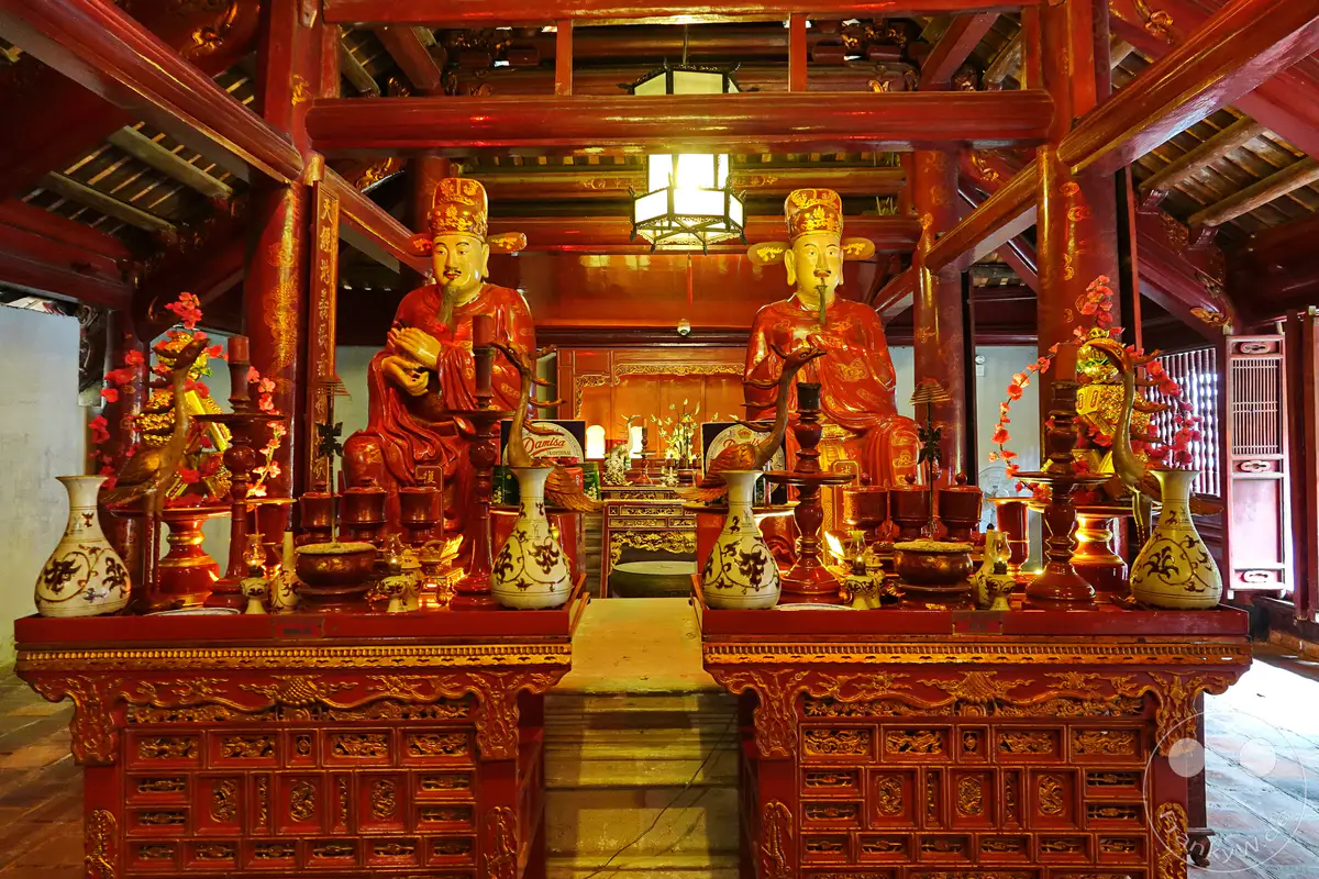 Im Inneren des Literaturtempels in Hanoi stehen zwei prächtige, rot und goldfarbene Statuen auf Altären, flankiert von Vasen und Opfergaben. Die aufwendig geschnitzten Holzstrukturen und die festliche Beleuchtung schaffen eine ehrwürdige und spirituelle Atmosphäre. Über den Statuen erstreckt sich die beeindruckende Holzkonstruktion des Tempeldaches, verziert mit traditionellen Ornamenten.
