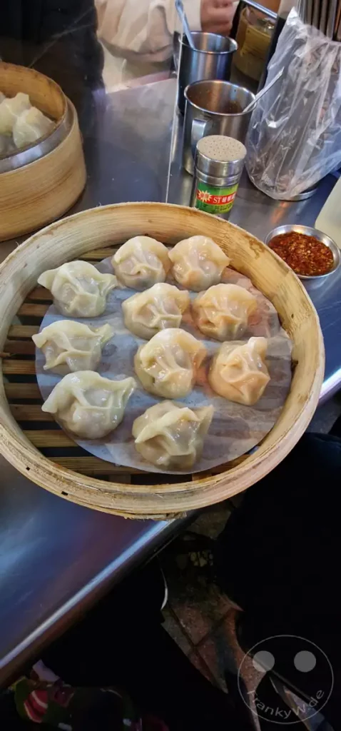 Eine Dampfkorbschale voller dampfender Teigtaschen, serviert auf einem Tisch in Taipeh, Taiwan. In der Nähe stehen Gewürzstreuer und eine kleine Schale mit Chiliflocken, was auf ein lebhaftes Streetfood-Erlebnis auf dem Ningxia Nightmarket hindeutet. Die warme Beleuchtung und die Nahaufnahme der Speisen erzeugen eine authentische und einladende Atmosphäre.