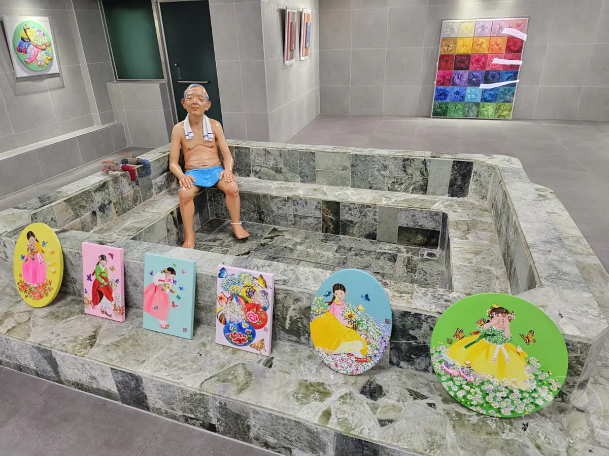 Südkorea - Busan - Culture Village Gamcheon - Eine interaktive Kunstinstallation mit einer Skulptur eines älteren Mannes, der in einem traditionellen Badehaus sitzt, umgeben von bunten Gemälden mit Kindern in Hanbok. Die Szenerie kombiniert Elemente der koreanischen Kultur und modernen Kunst in einer humorvollen Darstellung.