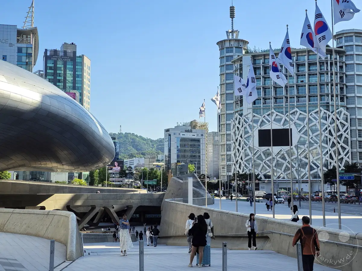 In Seoul, Südkorea, erhebt sich der futuristische Dongdaemun Design Plaza (DDP) vor einer Kulisse moderner Gebäude und einer bewaldeten Hügelkette. Koreanische Flaggen wehen im Wind und unterstreichen die nationale Identität des urbanen Zentrums. Die Szene vermittelt eine lebendige Atmosphäre, die historische und zukunftsweisende Elemente vereint.
