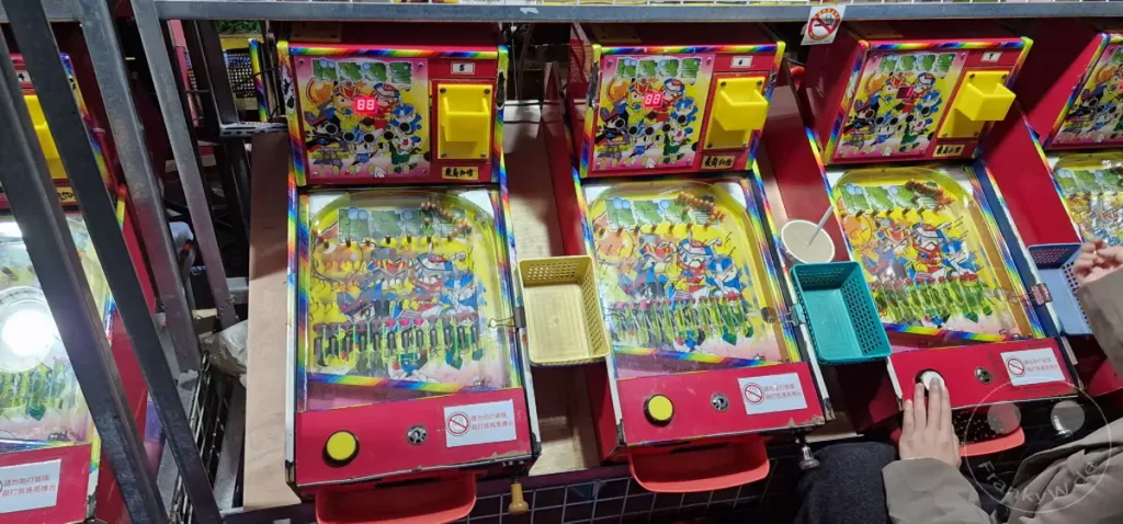 Mehrere bunte Pachinko-Spielautomaten stehen nebeneinander in einer Spielhalle im Ningxia Nightmarket in Taipeh, Taiwan. Die Automaten sind mit japanischen Charakteren und bunten Lichtern dekoriert, was eine lebhafte und unterhaltsame Atmosphäre schafft. Mehrere Körbchen für Kugeln und Münzschlitze sind zu sehen, während einige Personen spielen oder auf ihren Einsatz warten.
