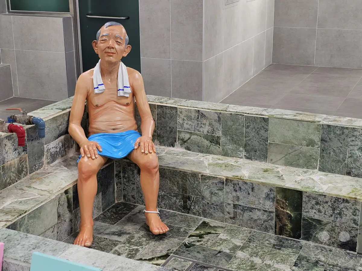 Eine lebensechte Skulptur eines älteren Mannes mit einem Handtuch um den Hals sitzt in einem traditionellen koreanischen Badehaus. Die Szene vermittelt eine Atmosphäre der Entspannung und kulturellen Immersion in Südkorea, genauer gesagt im Culture Village Gamcheon in Busan. Die detaillierte Darstellung der Figur und der Umgebung lädt den Betrachter ein, in die koreanische Badekultur einzutauchen.