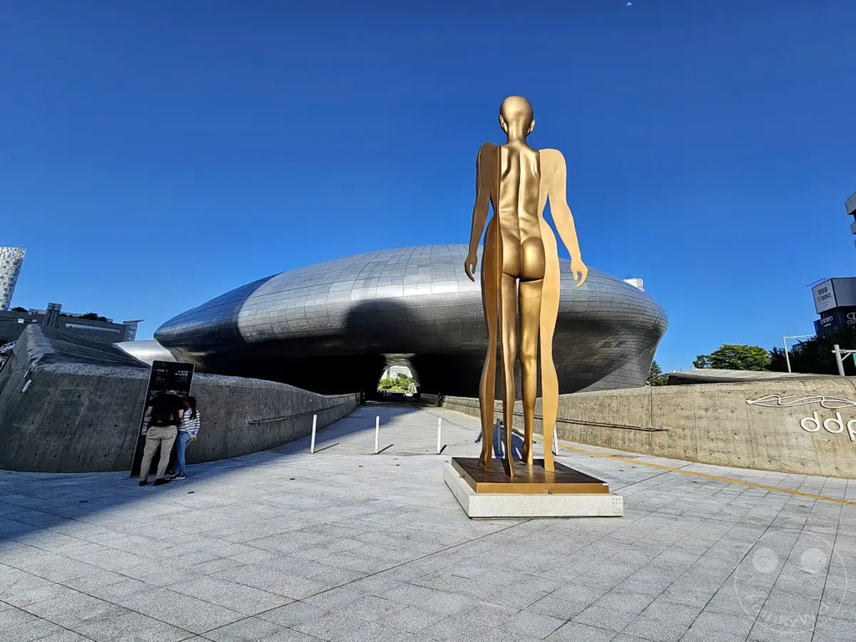 Südkorea – Seoul – Das Dongdaemun Design Plaza (DDP) wird von einer markanten goldenen Skulptur dominiert, die eine menschliche Figur darstellt. Die futuristische Architektur des DDP mit seiner glatten, metallischen Oberfläche bildet einen beeindruckenden Kontrast zur klaren blauen Himmelskulisse. Die Szene vermittelt eine Atmosphäre der modernen urbanen Ästhetik und des kulturellen Ausdrucks.