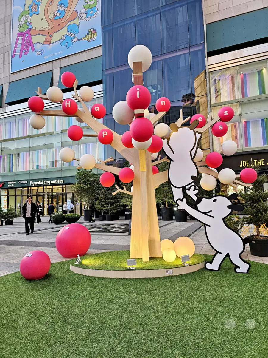 Eine dekorative Installation eines Baumes mit bunten Kugeln und Buchstaben, die das Wort "happiness" bilden, schmückt den Außenbereich der Hyundai City Outlets in Seoul, Südkorea. Zwei Comicfiguren scheinen mit dem Baum zu interagieren und tragen zur festlichen und verspielten Atmosphäre bei. Helles künstliches Licht beleuchtet die Szene und hebt die Details der Dekoration hervor.