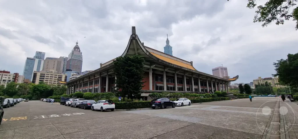 Die Sun-Yat-sen-Gedächtnishalle in Taipeh, Taiwan, präsentiert sich unter einem dramatischen Wolkenhimmel, der dem monumentalen Gebäude eine besondere Atmosphäre verleiht. Davor reihen sich Fahrzeuge auf einem weitläufigen Platz, während im Hintergrund moderne Hochhäuser die Skyline der Metropole prägen. Die Szene kombiniert traditionelle asiatische Architektur mit dem geschäftigen Treiben einer modernen Großstadt.