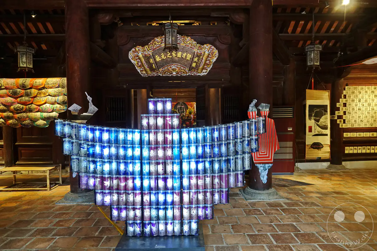 Im Tempel der Literatur in Hanoi, Vietnam, leuchtet eine beeindruckende Kunstinstallation aus zahlreichen zylindrischen Laternen in Blau- und Violetttönen. Diese Installation bildet eine Art architektonisches Element und steht im Kontrast zur traditionellen Holzarchitektur des Gebäudes. Die Szene strahlt eine Atmosphäre der kulturellen Verbindung zwischen Tradition und moderner Kunst aus.