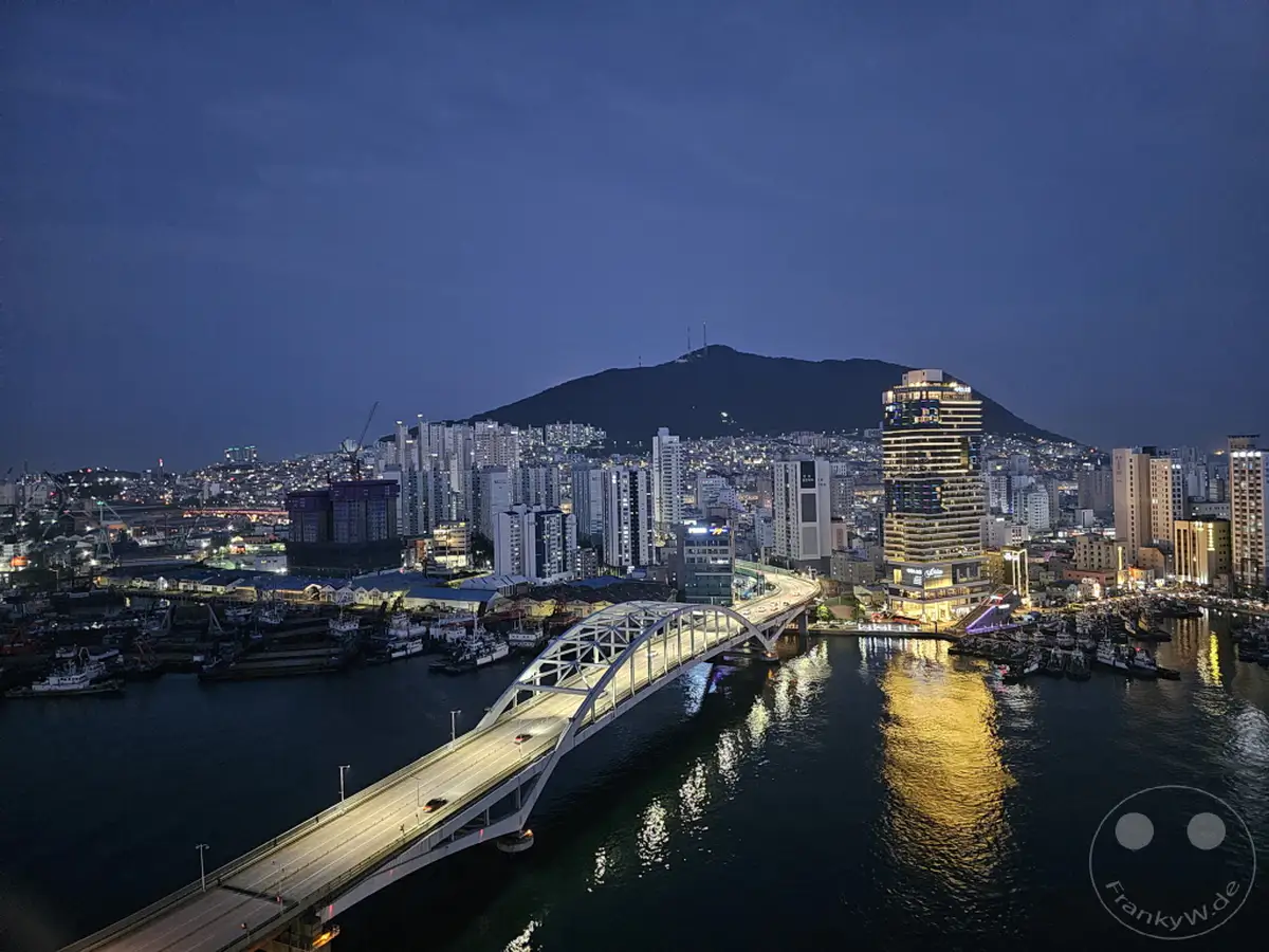 Ein weitläufiger Blick über die Stadt Busan in Südkorea bei Nacht offenbart eine beleuchtete Skyline mit einem markanten Wolkenkratzer und einer breiten Brücke, die sich über das dunkle Wasser spannt. Am Ufer liegen zahlreiche Boote und Schiffe in einem geschäftigen Hafen, während der Nachthimmel sich tiefblau über dem beleuchteten Panorama wölbt. Die Szene strahlt eine ruhige, aber lebendige Atmosphäre der Stadt aus, die von zahlreichen Lichtern erhellt wird.