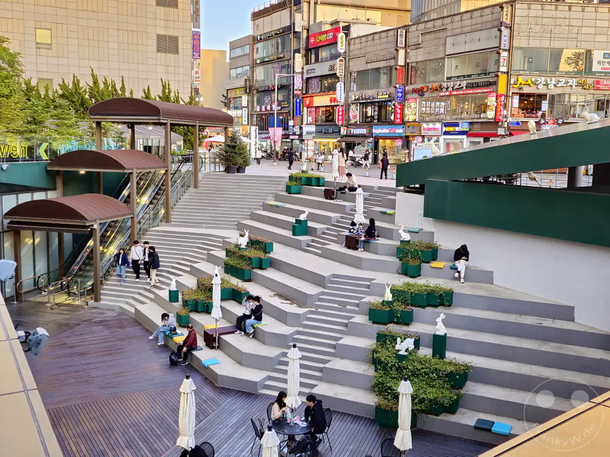 Eine lebhafte Treppenanlage im Außenbereich der Hyundai City Outlets in Seoul, Südkorea, lädt zum Verweilen ein. Menschen sitzen entspannt auf den Stufen und genießen die Atmosphäre zwischen Geschäften und Grünflächen. Mehrere Dekorationen in Form von Hasen zieren die Anlage und verleihen ihr einen verspielten Charakter.